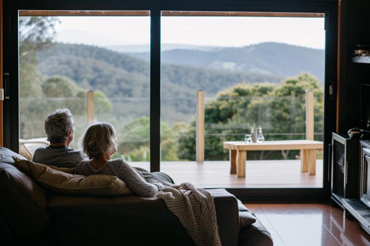 A retired couple enjopying the view from their picture window looking across a mountainscape.
