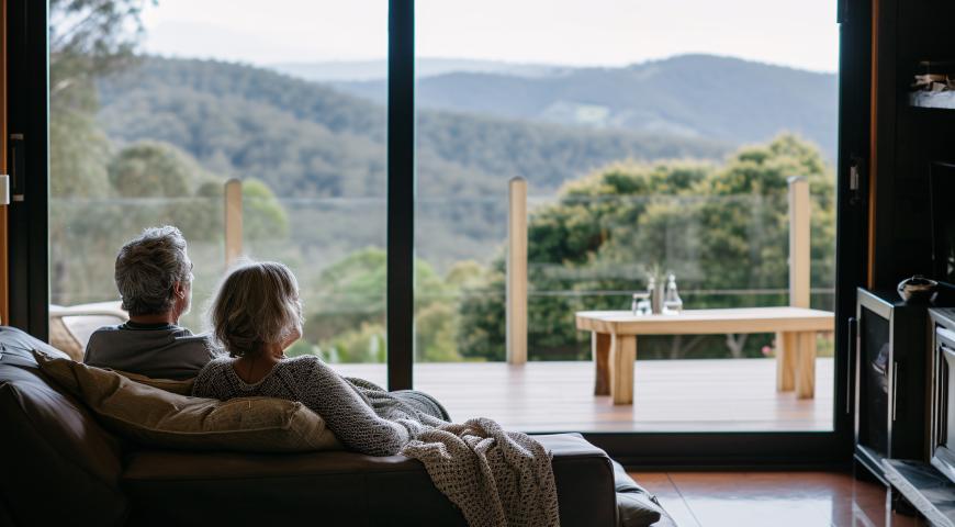 A retired couple enjopying the view from their picture window looking across a mountainscape.